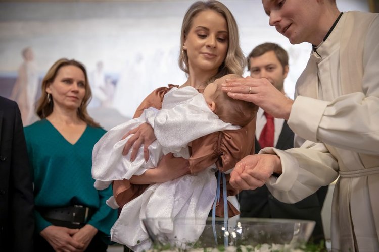 nainen pitää sylissä vauvaa ristiäisissä jota pappi kastaa