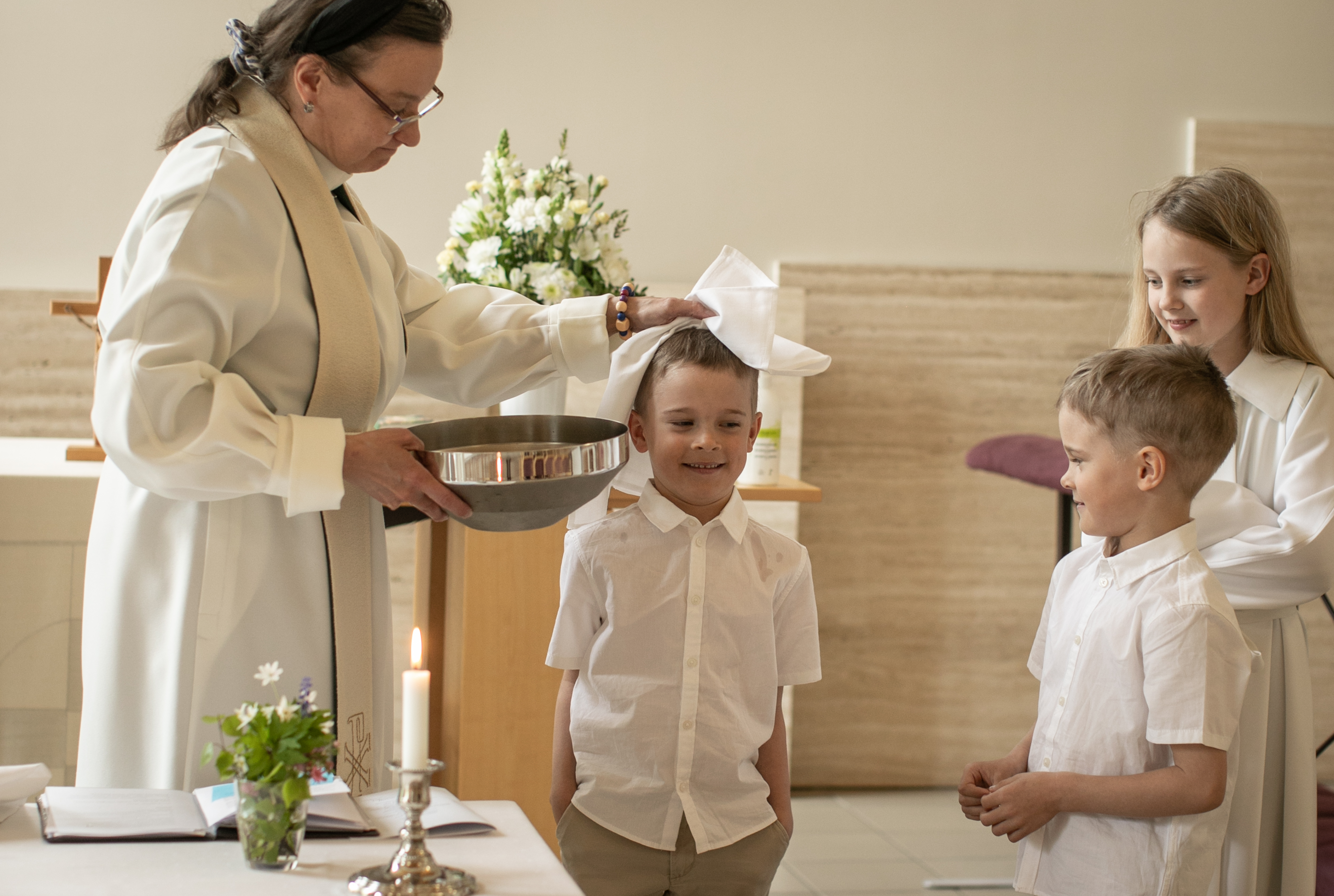 Kuvassa on pappi ja kolme lasta. Pappi kuivaa kastetun lapsen päätä valkoisella liinalla. Kaikki hymyilevät.