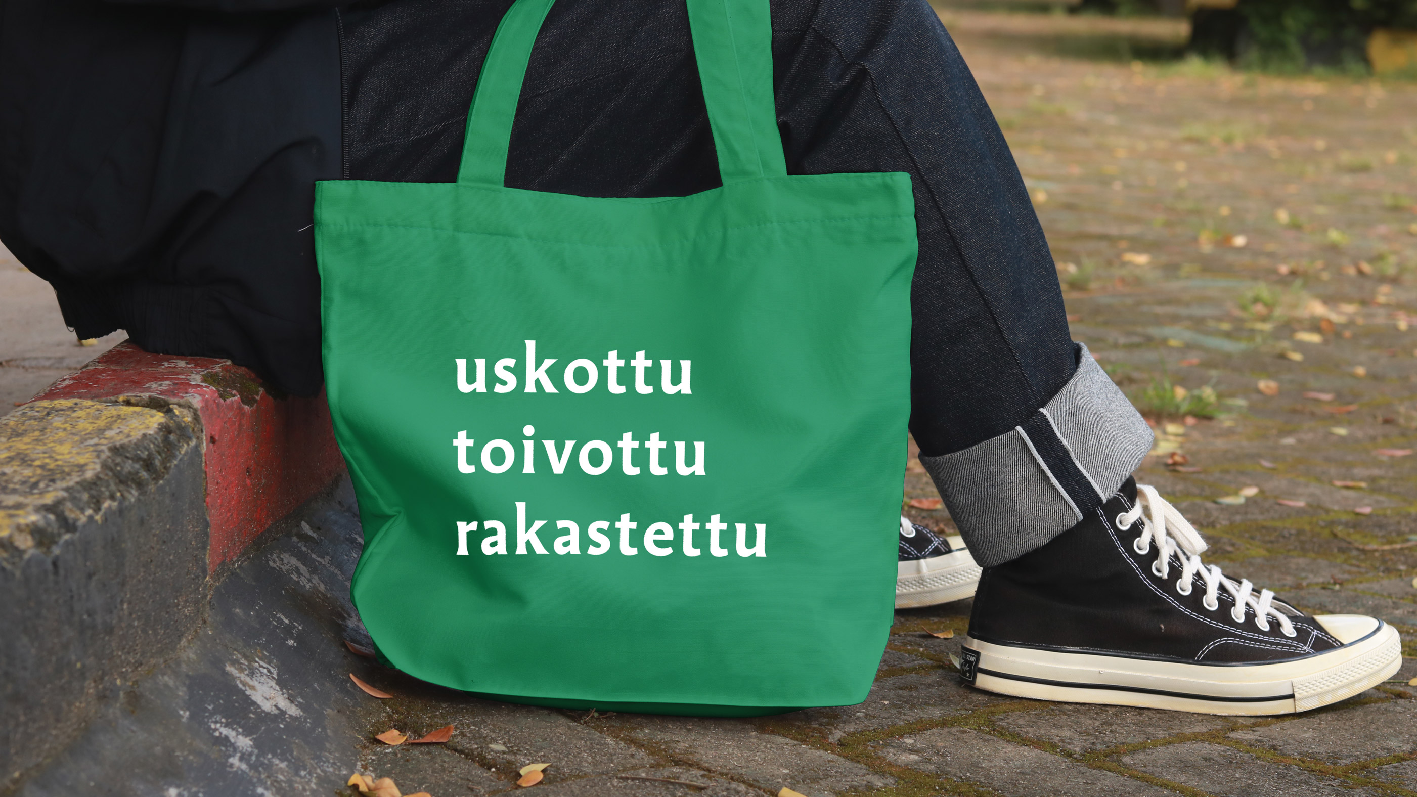 Kiveyksellä istuva henkilö kuvattuna alavartalon osalta. Jalassa tennarit ja vieressä kassi, jossa on tekst...
