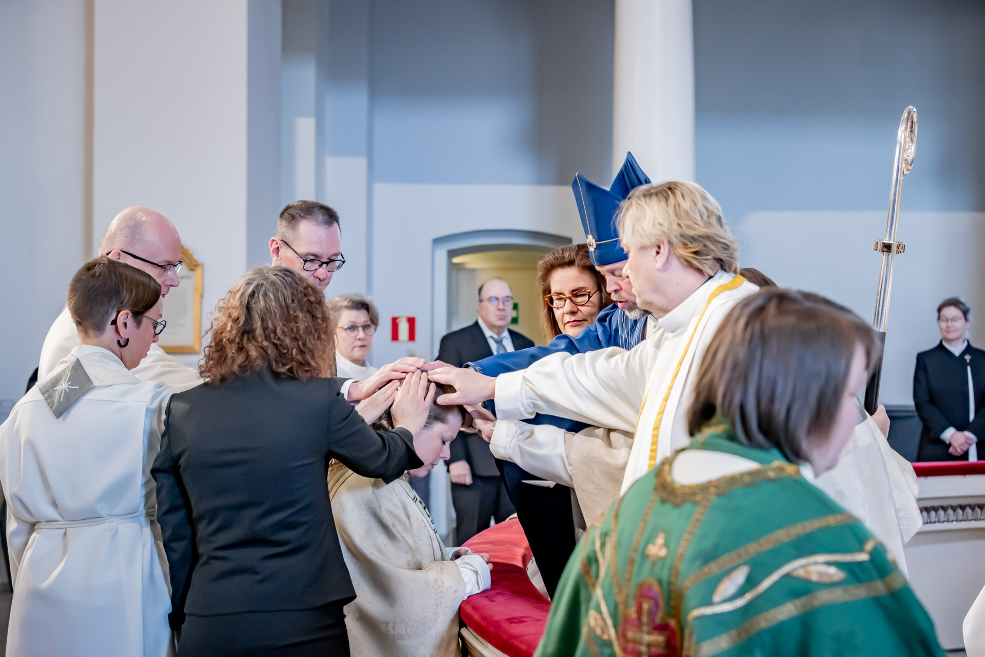 Ihmisiä siunaamassa tulevaa pappia alttarilla, juhlavaa.
