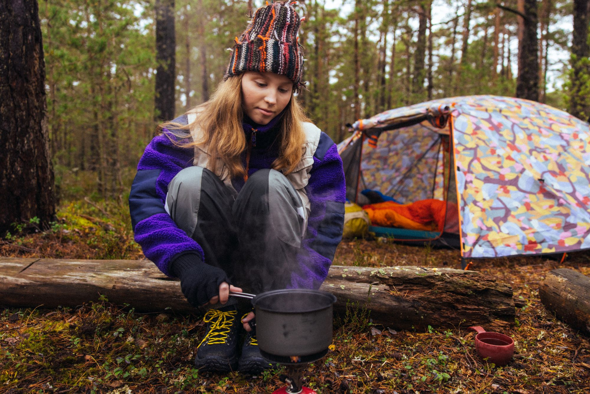 Nuori nainen, partio, kokkaa telttansa edustalla metsässä.
