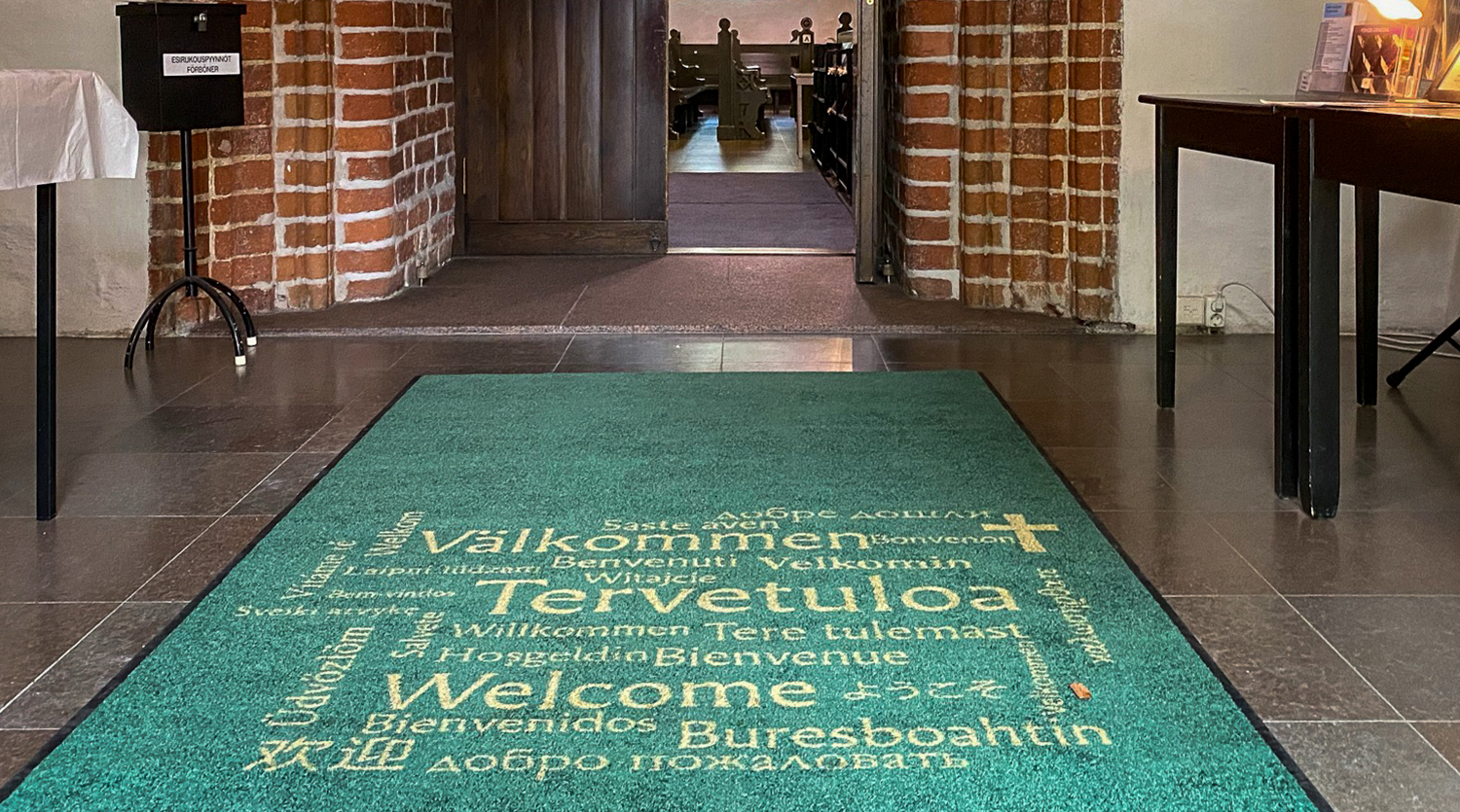 Eteinen kirkossa jossa vihreä matto tekstillä Tervetuloa monella kielillä.